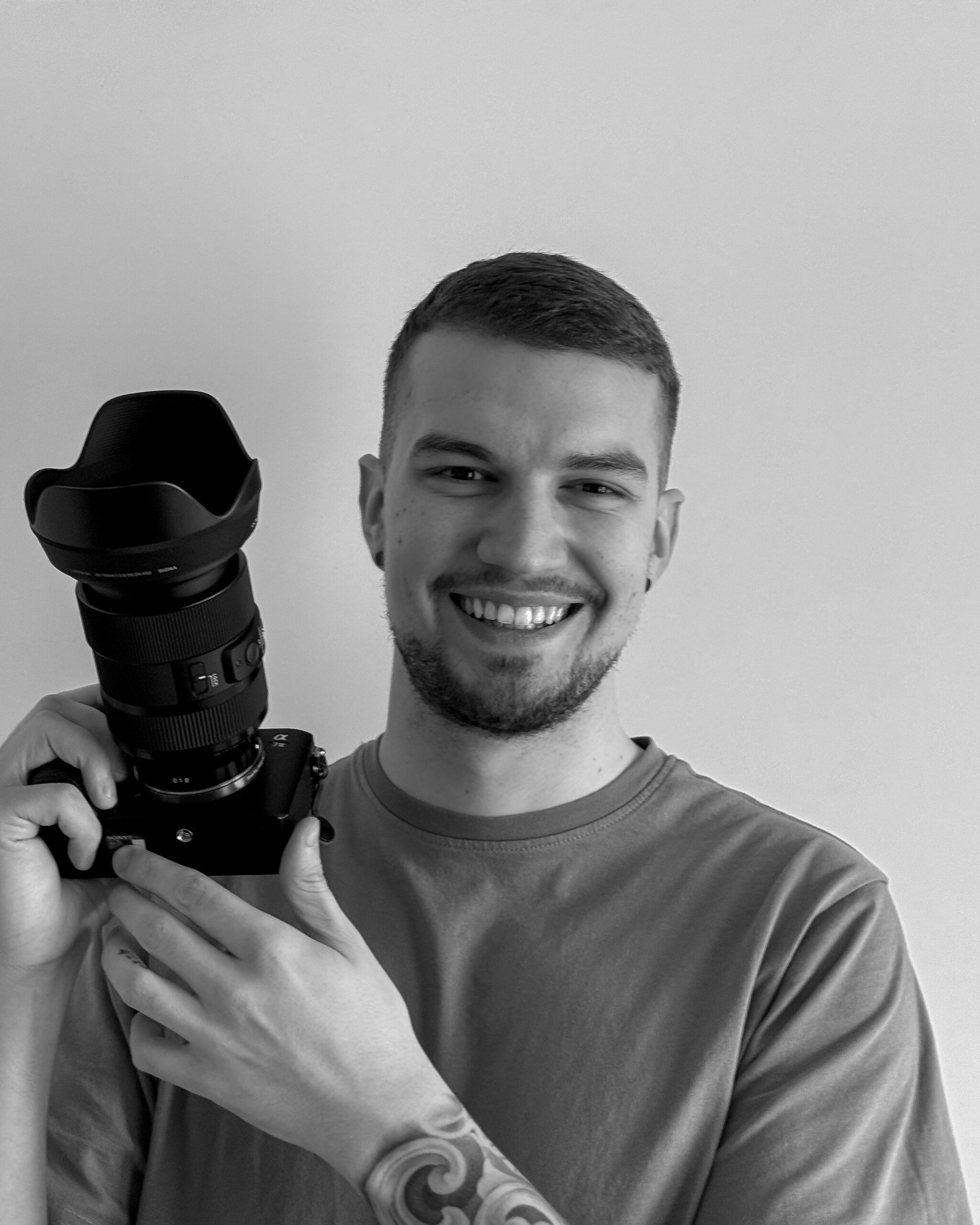 Black and white portrait of Rovinj photographer Dominick Lutrovic smiling while holding a camera with a large lens.